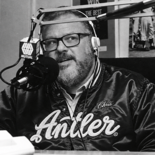 Black-and-white portrait of Chris Stewart wearing headphones and an Antler jacket at a radio microphone.